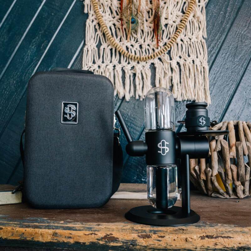 Stündenglass Kompact Gravity Infuser in black with carrying case, set against a rustic wooden backdrop.