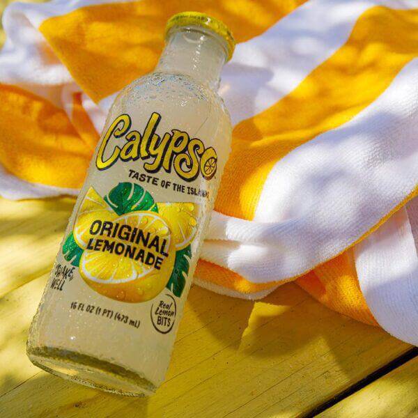 Calypso Lemonade 16oz bottle on a wooden table with a vibrant orange and white beach towel.