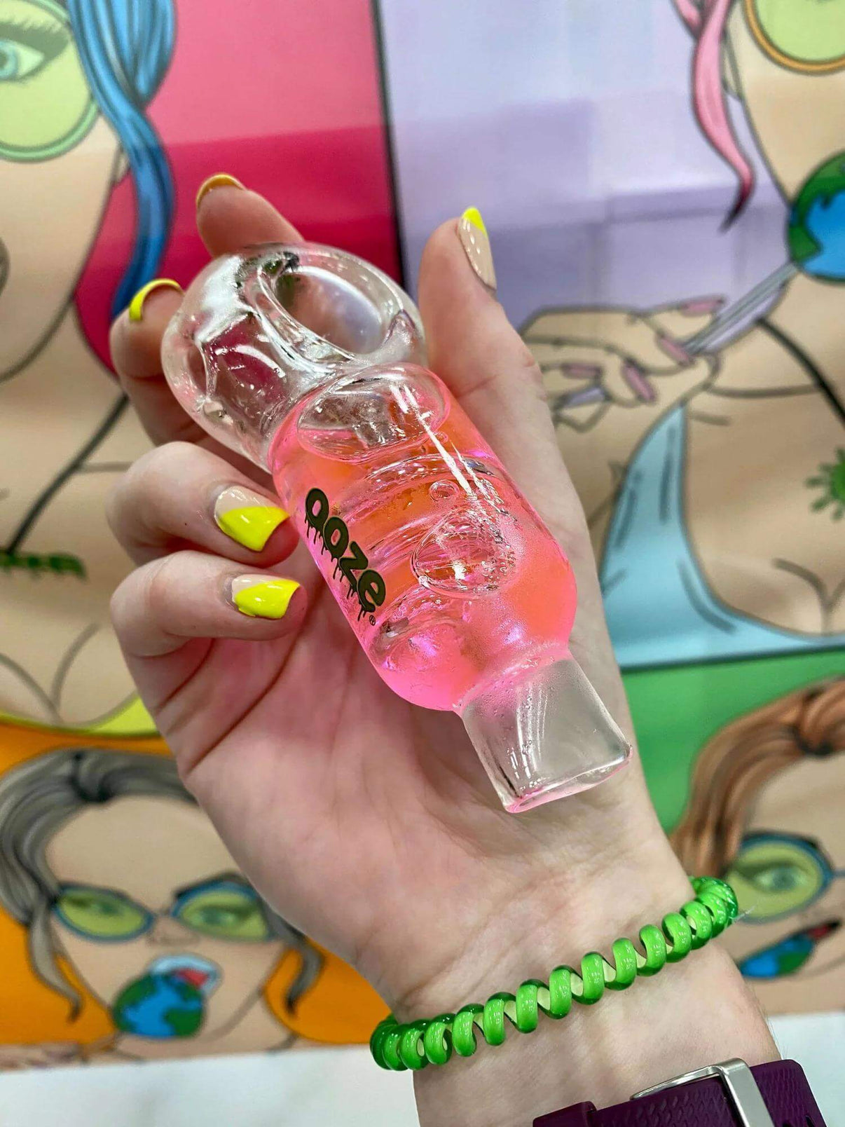 Hand holding Ooze Cryo Freezable Glycerin Glass Hand Pipe with pink glycerin, showcased in a colorful backdrop.