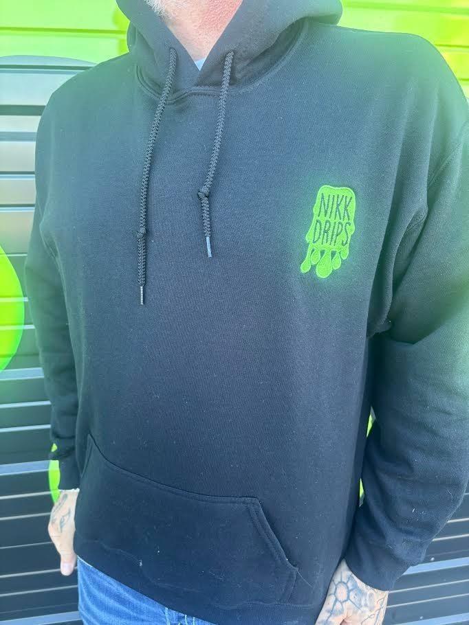 Close-up of a person wearing a Nikk Drips Hoodie displaying its green logo on a black fabric backdrop.