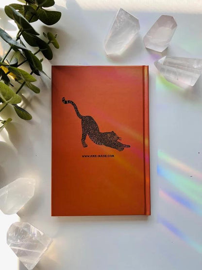 Ark Made Leaping Leopards Lined Journal with orange cover and playful leopard design, surrounded by crystals and greenery.