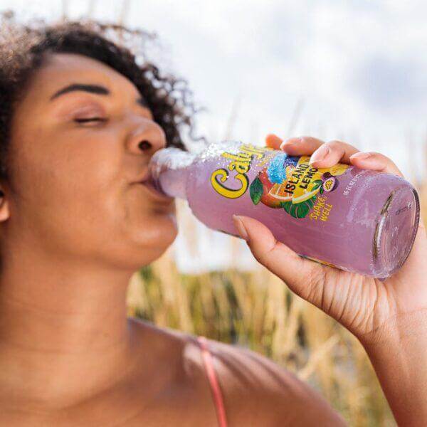 Woman enjoying Calypso Lemonade 16oz bottle with vibrant fruit flavors outdoors.