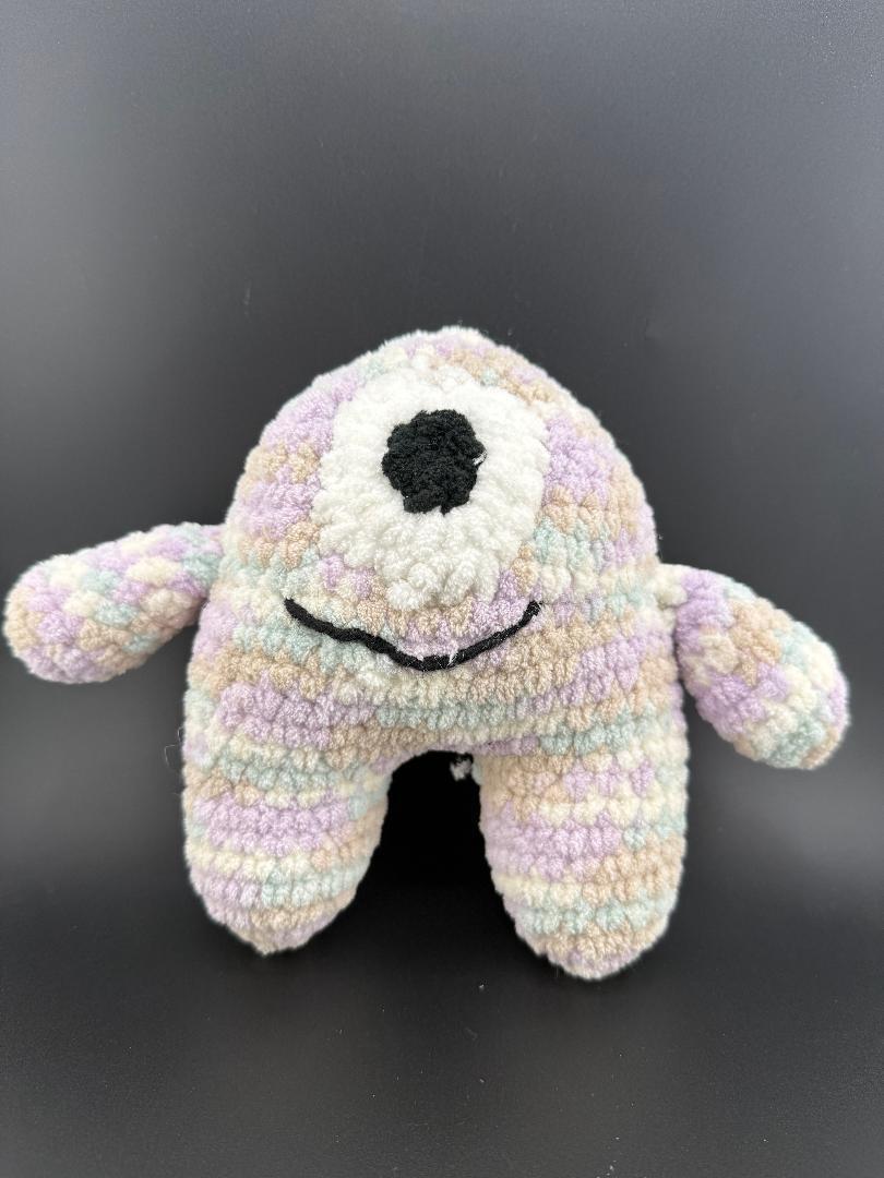 Crochet one-eyed monster plush toy in pastel colors, featuring a black smile and soft, fuzzy texture.