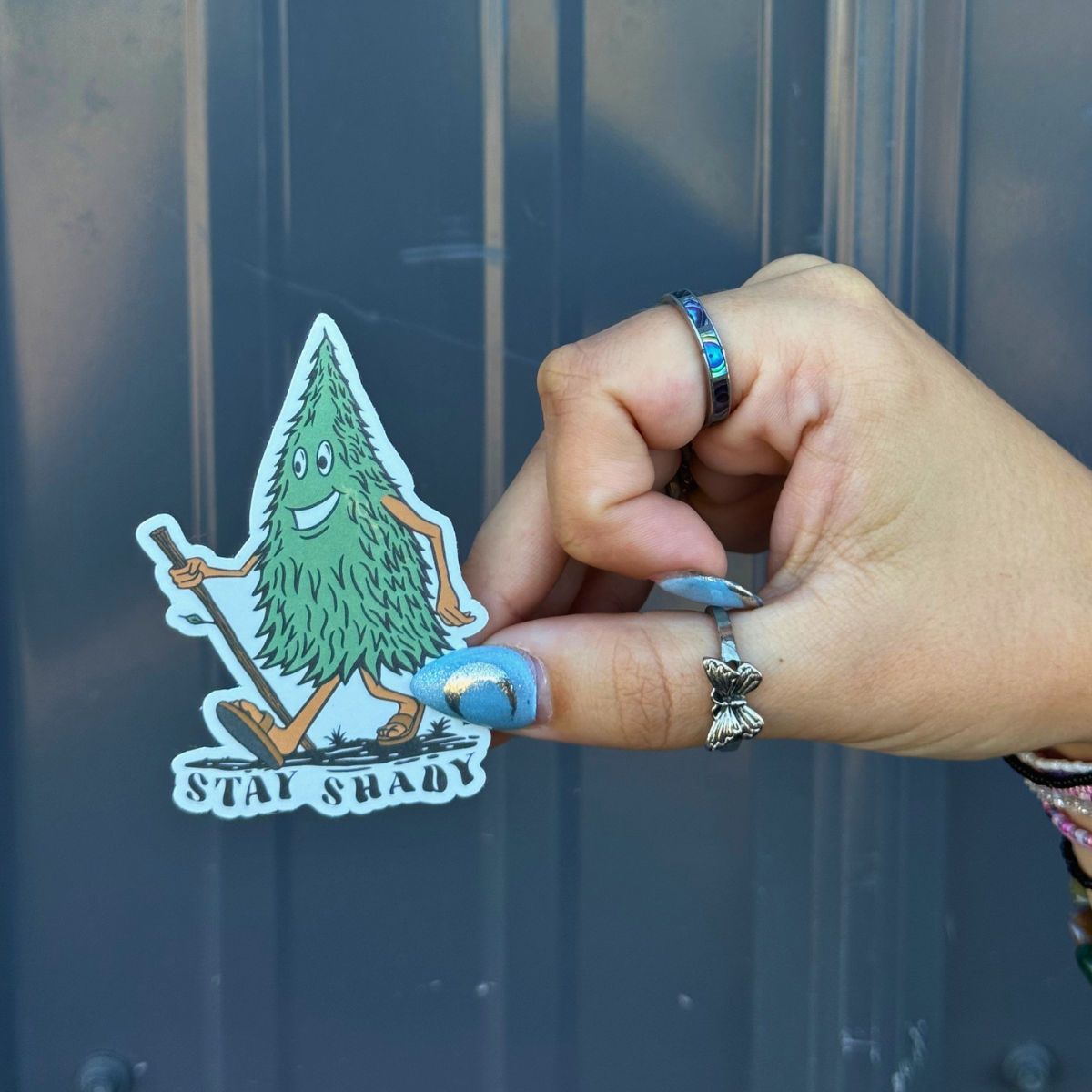 Hand holding a BBB Sticker | Stay Shady featuring a cartoon tree character outdoors.