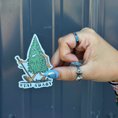 Hand holding a BBB Sticker | Stay Shady featuring a cartoon tree character outdoors.