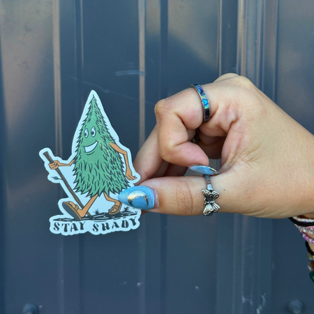 Hand holding a BBB Sticker | Stay Shady featuring a cartoon tree character outdoors.