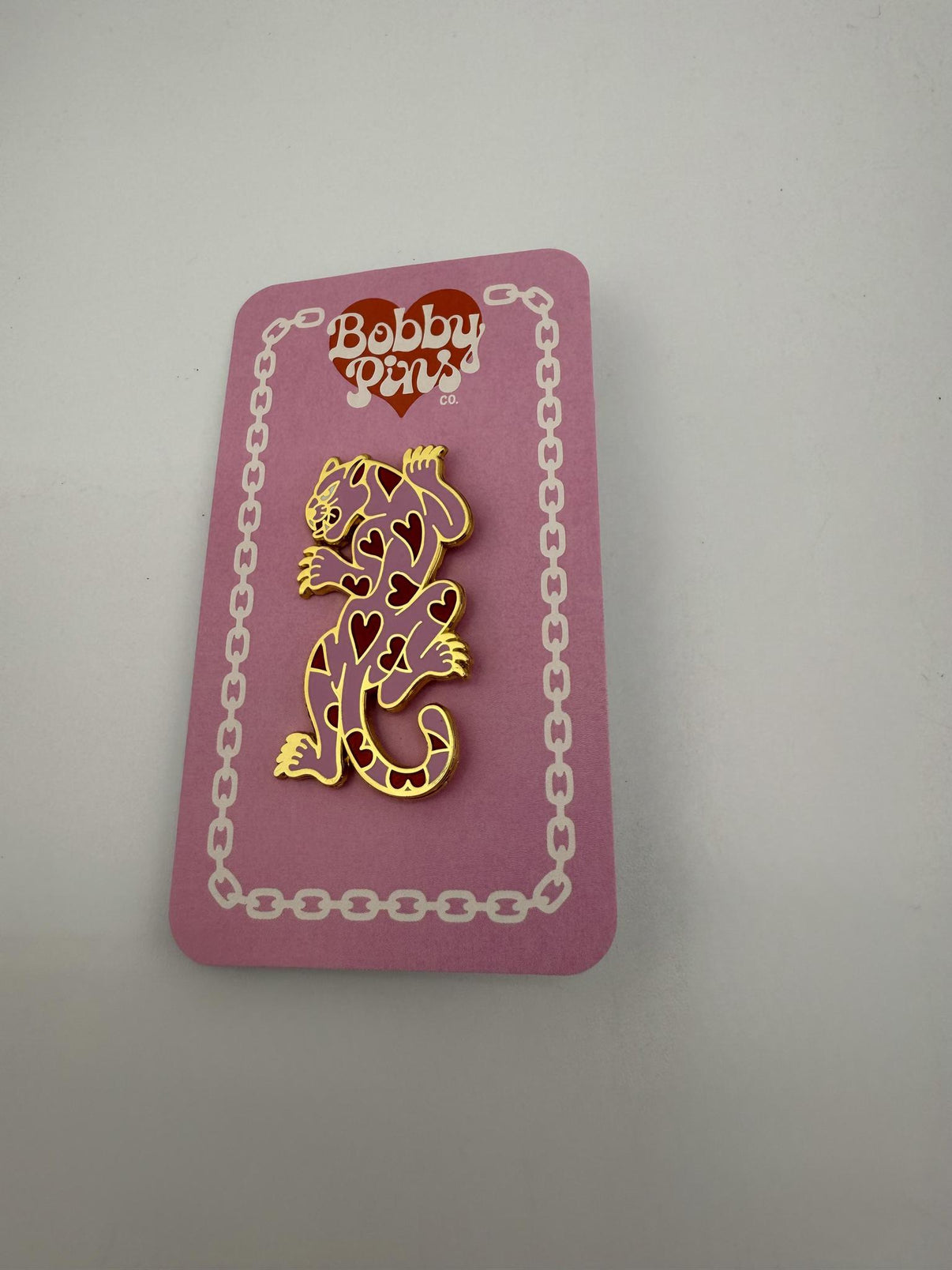 Bobby Pins Co. Enamel Pin - Heart Panther displayed on a pink backing card, featuring a gold-colored heart pattern.