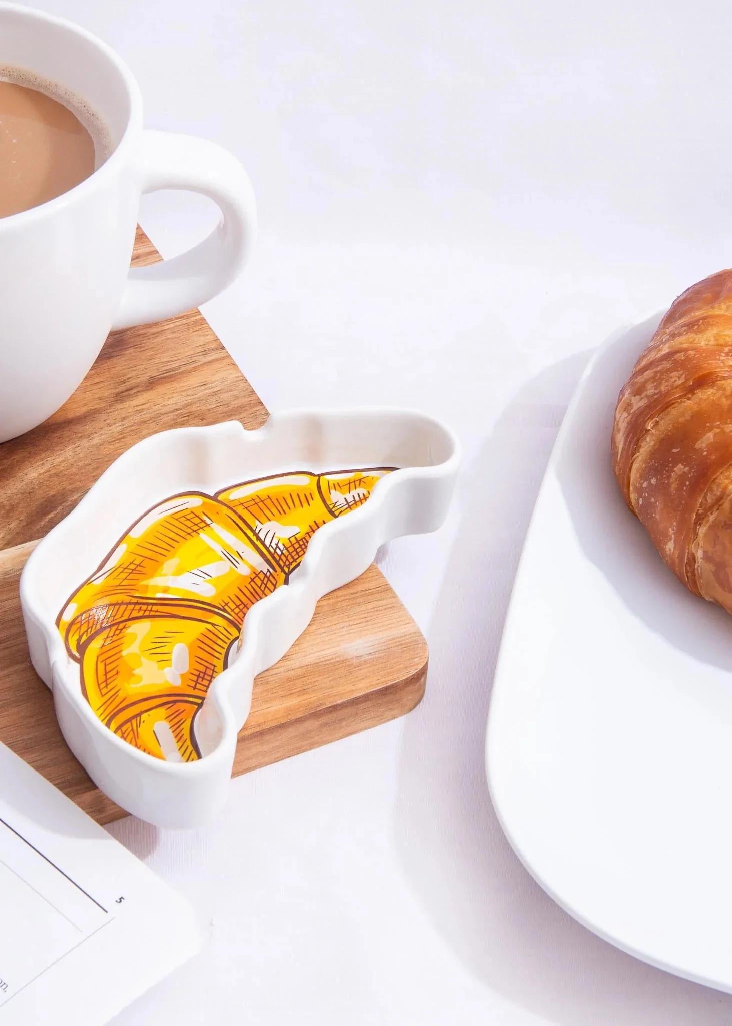 Canna Style Ashtray - Croissant displayed beside a cup of coffee and a real croissant on a wooden surface.