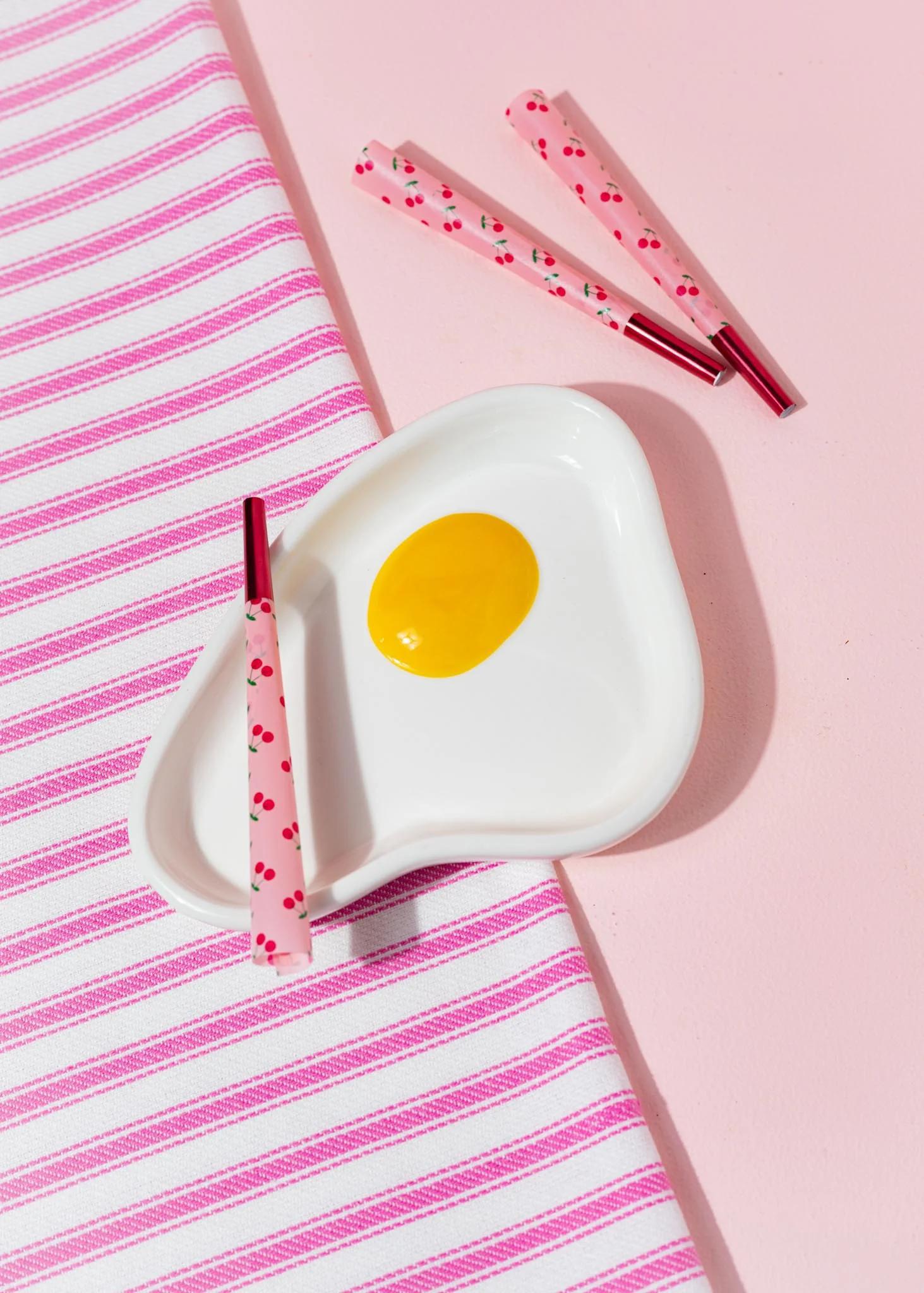 Canna Style Ashtray - Egg, whimsical egg-shaped ashtray with pink smoking accessories on striped fabric.