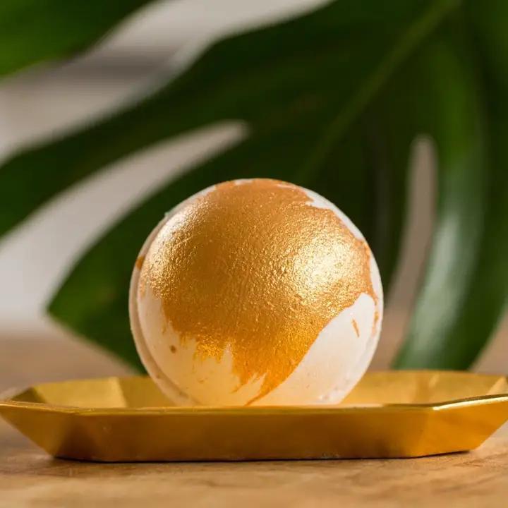 Whipped Up Wonderful Cleopatra Milk And Honey Bath Bomb displayed on a golden tray with a leaf backdrop.