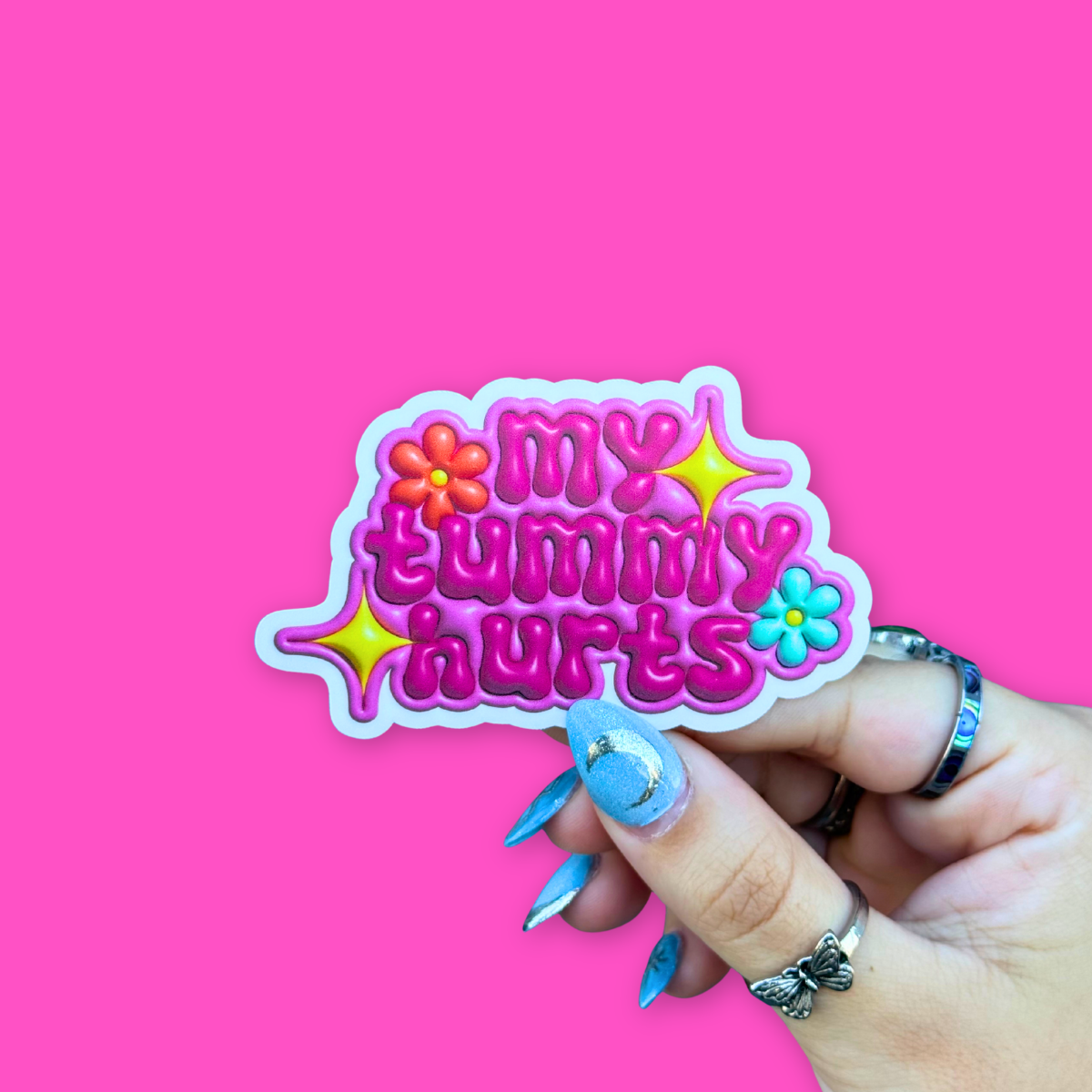 Hand holding a colorful BBB Sticker | My Tummy Hurts against a vibrant pink background.