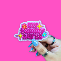 Hand holding a colorful BBB Sticker | My Tummy Hurts against a vibrant pink background.