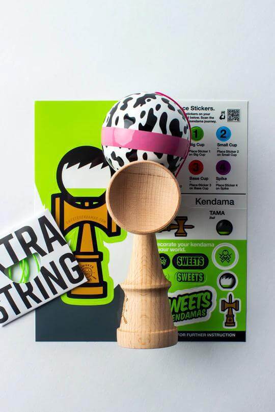 Sweets Kendama Mini with cow-patterned tama, extra string, and instructional stickers on colorful background.