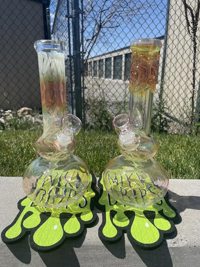 Two glass bongs with colorful designs and drip mats on grass, showcasing unique shapes and artisan craftsmanship.