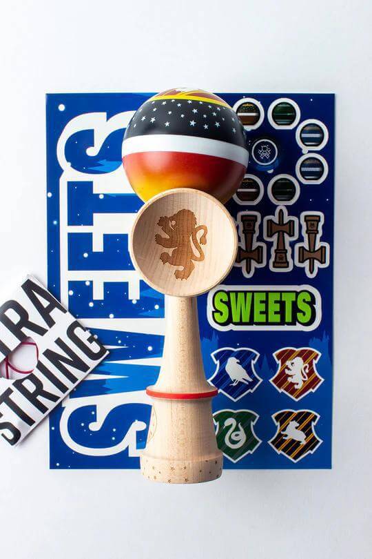 Sweets Kendama V35 in Sticky Clear with lion engraving, stickers, and extra string on a blue background.