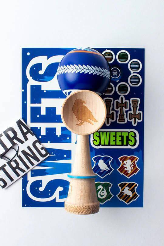 Sweets Kendama V35 Sticky Clear with engraved raven cup and colorful stickers on a vibrant blue background.