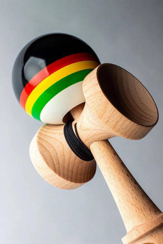 Sweets Kendama Triple Stripe with black, red, yellow, and green design on tama against a soft gray background.