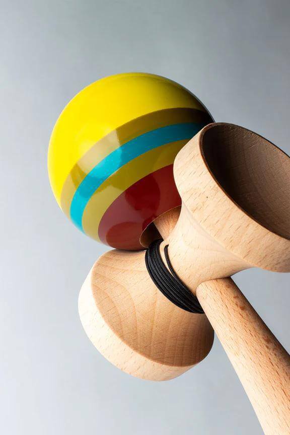 Sweets Kendama Triple Stripe, featuring vibrant yellow tama with blue and red stripes, and natural beech wood handle.