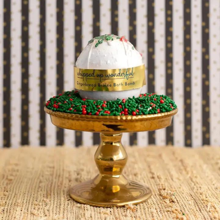 Whipped Up Wonderful Gingerbread Brulee Bath Bomb displayed on a gold cake stand with festive decorations.
