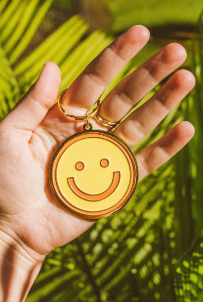 Wild Rose Keychain - Smiley held in a hand with palm trees in the background, featuring a cheerful smiley face design.