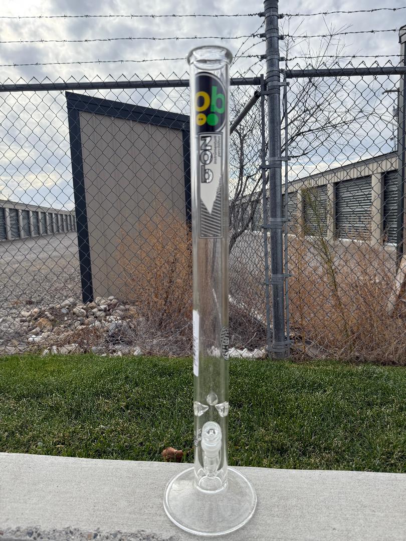 Zob Glass 24" Straight Tube Flower Piece standing outdoors against a chain-link fence with grass in foreground.